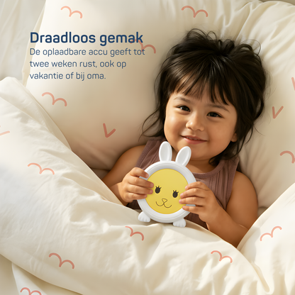 Kind ligt lachend in bed met een witte slaaptrainer in de vorm van een konijn , draadloos oplaadbaar model dat tot twee weken meegaat, ideaal voor thuis of op vakantie.
